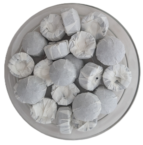 Draufsicht auf mehrere, kleine, kreisförmige Puer-Tee-Mini-Fladen, die einzeln in weißes, gefaltetes Papier eingewickelt sind und auf einem klaren Glasteller liegen.