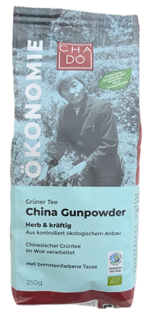 Cha Dô Ökonomie Bio China Gunpowder Grüner Tee Verpackung in Hellblau mit Foto einer asiatischen Tee-Anbauerin. Bildbeschreibung: Verpackung mit hellblauem Etikett, Schwarz-Weiß-Foto einer asiatischen Frau im Teegarten und grünen Bio- sowie Fairtrade-Logo