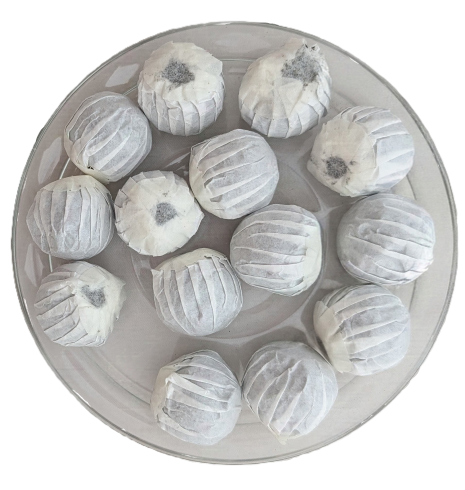 Draufsicht auf eine runde, durchsichtige Glasschale, in der etwa zwölf kleine Pu-Erh-Tee-Drachenperlen liegen. Jede Kugel ist individuell in dünne, gefaltete weiße Papiereingewickelt. Bei einigen Perlen schimmert der dunkle Tee an der oberen Öffnung durch